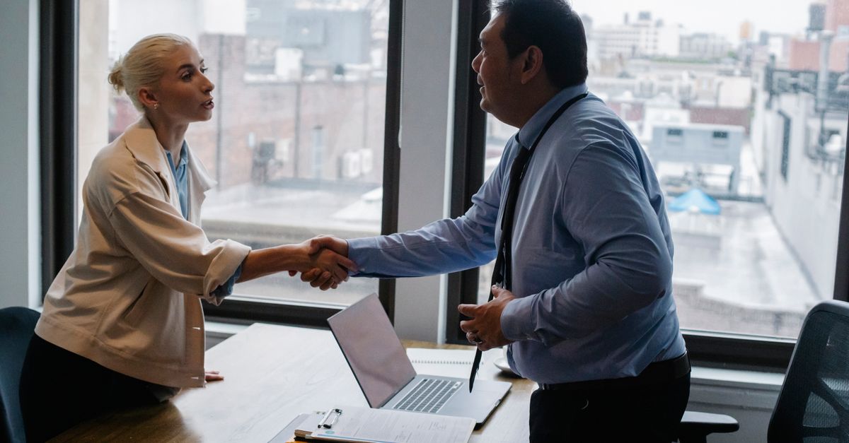 Young female candidate shaking hands with employer before job interview in light office with urban landscape through windows